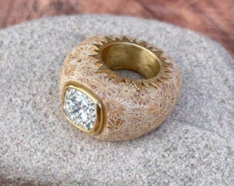 May include: A unique ring featuring a large, square-cut diamond set in a gold bezel. The ring's band is a light beige color with a textured pattern, and the top edge has a gold, saw-tooth design.