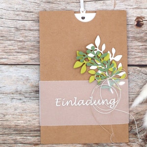May include: A brown paper card with a white string attached to the top. The card has a clear plastic window with the word "Einladung" printed in white. The card is decorated with green and white paper leaves.
