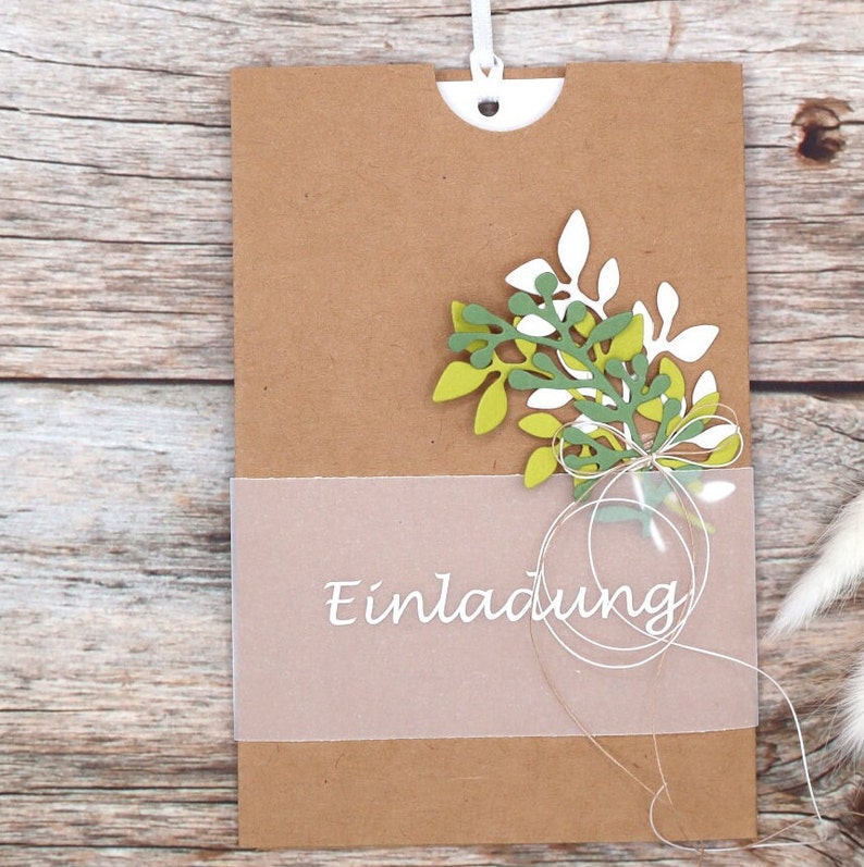 May include: A brown paper card with a white string attached to the top. The card has a clear plastic window with the word "Einladung" printed in white. The card is decorated with green and white paper leaves.
