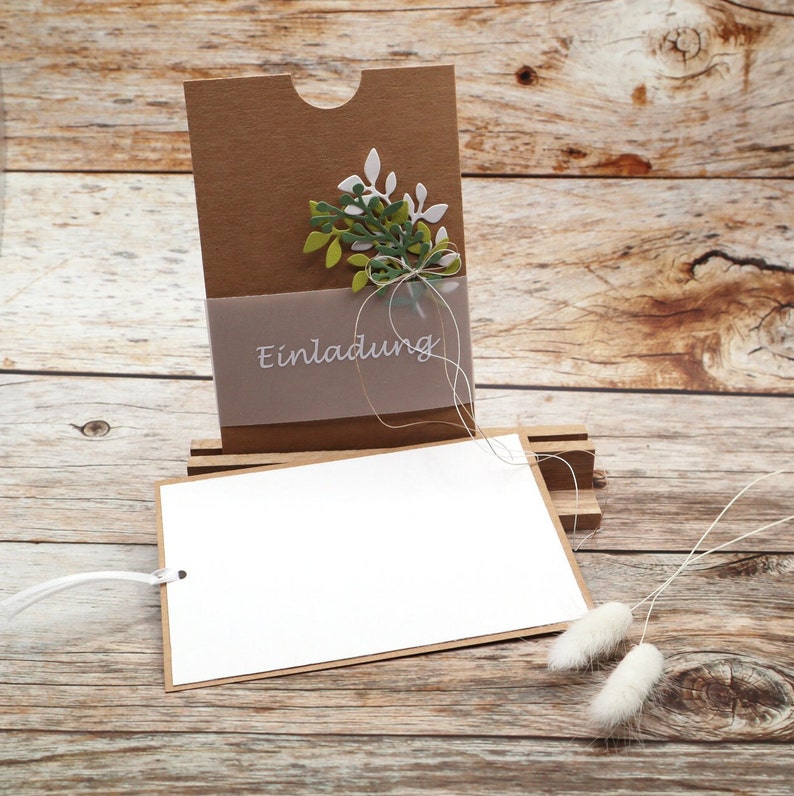 May include: A brown card with a cut-out slot and a green and white floral design. The card has a white label with the word "Einladung" printed on it. The card is sitting on a wooden surface.