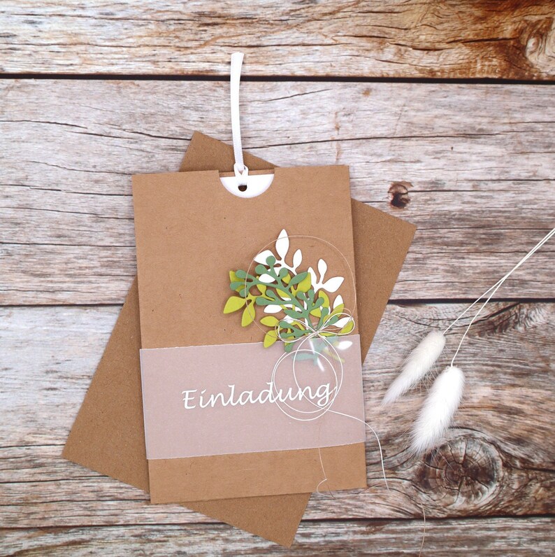 May include: A brown paper envelope with a white string attached to the top. The envelope has a clear plastic window with a green and white floral design and the word "Einladung" printed on it.