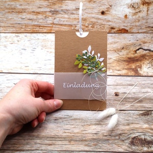 May include: A brown card with a white string attached to the top. The card has a cut-out at the top and a light brown envelope with the word "Einladung" printed on it. The card is decorated with green and white paper leaves.