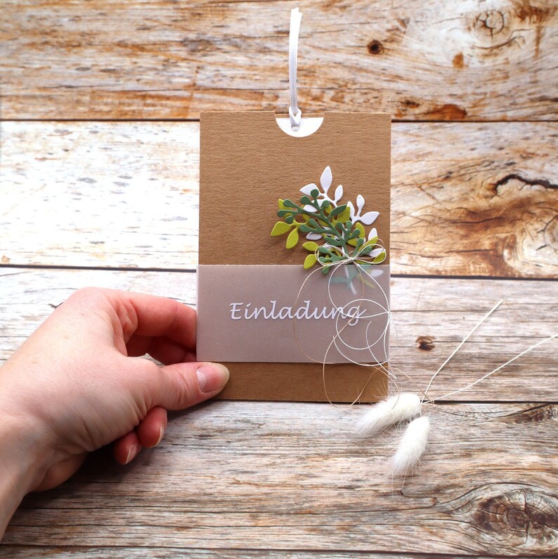 May include: A brown card with a white string attached to the top. The card has a cut-out at the top and a light brown envelope with the word "Einladung" printed on it. The card is decorated with green and white paper leaves.