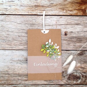 May include: A brown paper envelope with a white string attached to the top. The envelope has a white, translucent insert with the word "Einladung" printed on it. The envelope is decorated with a green and white paper flower.