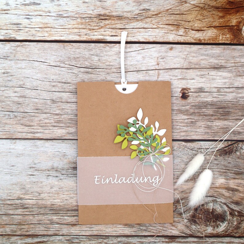 May include: A brown paper envelope with a white string attached to the top. The envelope has a white, translucent insert with the word "Einladung" printed on it. The envelope is decorated with a green and white paper flower.