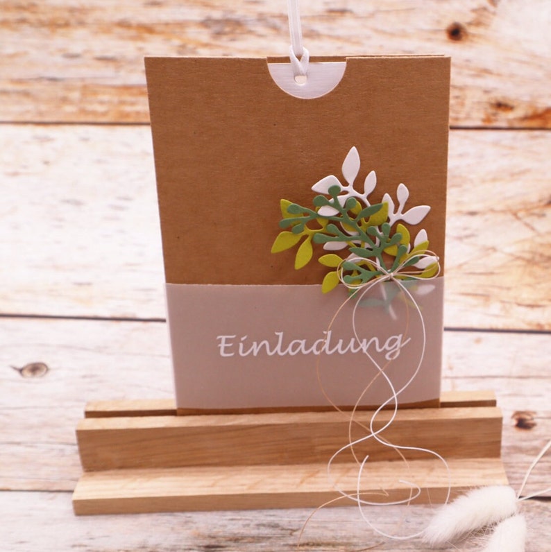 May include: A brown paper card with a white envelope attached to the top. The card has a white ribbon tied around it with a bow. The card has a green and white floral design and the word "Einladung" written on it.