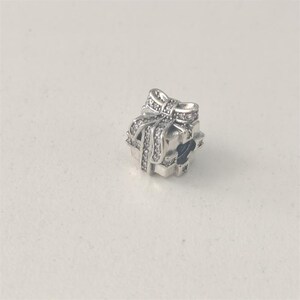 May include: A silver charm in the shape of a gift box with a bow on top. The charm is encrusted with small, clear gemstones. The gift box charm is a jewelry accessory.