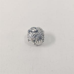 Pode incluir: Um berloque de prata com um design de concha, estrela do mar e caranguejo. O berloque apresenta texturas detalhadas e detalhes de contas, evocando um tema náutico. O berloque tem formato de cubo.