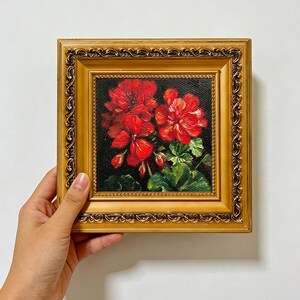 Può includere: Un dipinto a olio di vivaci fiori di geranio rosso con foglie verdi su sfondo scuro, racchiuso in una cornice quadrata decorata color oro metallizzato con intricati dettagli in rilievo lungo i bordi esterni della cornice.