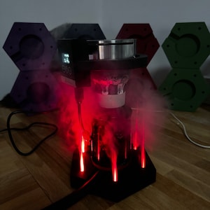 May include: A black and silver electric vaporiser with a digital display and red lights. The vaporiser is surrounded by smoke and is sitting on a wooden surface.