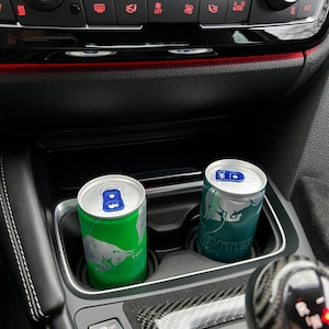 Könnte beinhalten: Zwei Dosen Red Bull Energy Drink, eine grüne und eine blaue, in einem Getränkehalter im Auto. Die grüne Dose hat den Text "The Red Bull" darauf. Die blaue Dose hat den Text "Winter Edition" darauf.