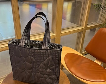 Könnte beinhalten: Eine gesteppte schwarze Tragetasche mit Reißverschluss und zwei Henkeln oben. Die Tasche steht auf einem Holztisch vor einem Fenster mit bunten Glasscheiben.