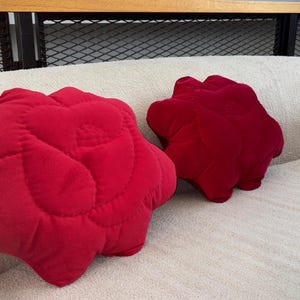 Puede incluir: Dos cojines decorativos rojos y afelpados con forma de flor descansan sobre una cama color crema. Los cojines tienen un diseño acolchado y son de un tono rojo vibrante, añadiendo un toque de color a la ropa de cama neutra.