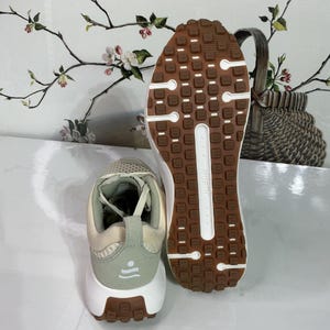 May include: A pair of athletic shoes with a beige and light green upper, white midsole, and brown outsole. The outsole features a textured pattern for grip. The heel has a light green accent with a small design. The shoes are designed for running or walking.