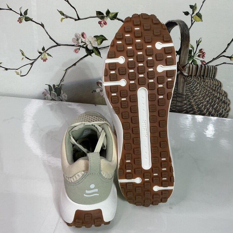 May include: A pair of athletic shoes with a beige and light green upper, white midsole, and brown outsole. The outsole features a textured pattern for grip. The heel has a light green accent with a small design. The shoes are designed for running or walking.