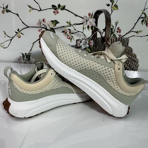 May include: Pair of athletic shoes in a neutral colour palette. The trainers feature a light green mesh upper with beige accents, white soles, and brown rubber outsoles. The shoes are designed for comfort and performance.