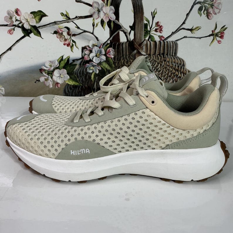 May include: A pair of beige and green athletic shoes with white soles and brown treads. The shoes feature a mesh upper and the word "HILMA" is printed on the side. Designed for running or other athletic activities.