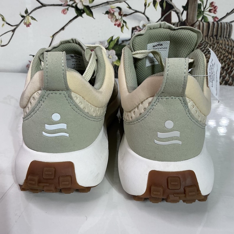 May include: A pair of athletic shoes in light green and beige. The shoes have a white sole with a brown tread. The heel features a white graphic of a circle above two curved lines. The brand name "HILMA" is visible on the back of one shoe.