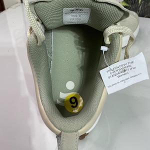 May include: An inside view of a beige and white trainer. The shoe features a green insole with a size 9 sticker. A tag reads "FROZEN DEW THE EVERYWHERE STANDARD IT (For Women)". The shoe is labeled "HILDA The Everywhere".
