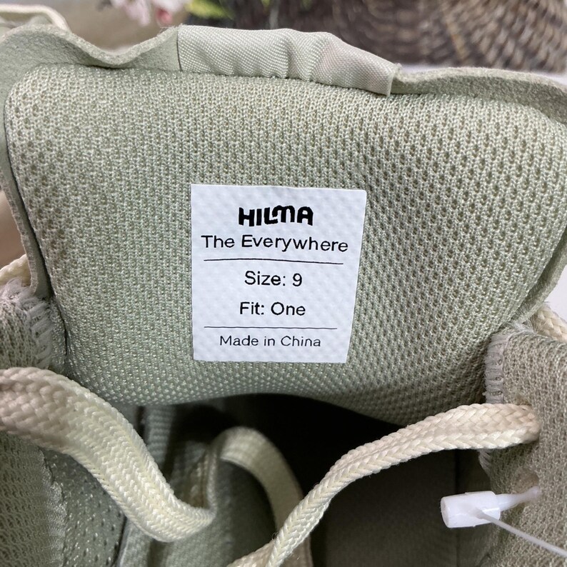 May include: Close-up of the inside of a pale green trainer. A white label reads "HILMA The Everywhere Size: 9 Fit: One Made in China". The laces are off-white.