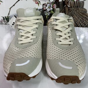 May include: A pair of beige athletic shoes, branded "HILMA" on the tongue. They feature a mesh upper, suede detailing, and brown soles. The design includes white laces and a curved white accent on the toe.