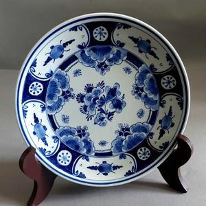 Könnte beinhalten: Ein weißer Keramikteller mit blauem Blumendesign. Der Teller hat ein zentrales Blumenbouquet und einen Rand mit stilisierten Blumen und geometrischen Formen. Der Teller wird auf einem Holzständer präsentiert.