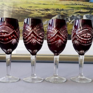 Può includere: Quattro calici da vino rosso rubino con steli e basi trasparenti. I calici presentano un design ornato e inciso attorno alla coppa. Lo sfondo presenta un dipinto di paesaggio incorniciato.