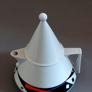 Peut inclure: Théière en céramique blanche avec un couvercle conique et un motif géométrique en noir, rouge et bleu.