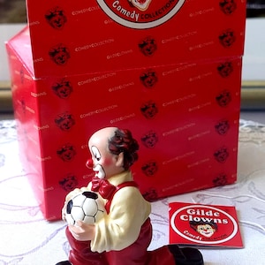 May include: A ceramic clown figurine from the "Gilde Clowns Comedy Collection" kneels, holding a soccer ball. The clown wears a red bow tie, red pants, and a cream-colored shirt. A red box with the same logo is behind the figurine.