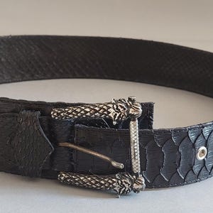 Peut inclure: Une ceinture noire avec une texture peau de serpent. La ceinture est ornée d'une boucle argentée au design décoratif et d'une boucle de ceinture assortie. La ceinture comporte plusieurs œillets pour le réglage.