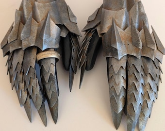 Könnte beinhalten: Ein Paar silberfarbene Metallhandschuhe mit schuppiger Textur und spitzen Spitzen. Die Handschuhe sind abgenutzt und haben eine abgenutzte Oberfläche. Ein Handschuh hat einen silbernen Ring mit einer Inschrift.