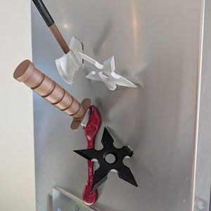 Puede incluir: Un refrigerador de acero inoxidable con tres objetos magnéticos adheridos a él. Los objetos son un hacha marrón y negro, un cuchillo marrón y negro y dos estrellas ninja negras. Las estrellas ninja están adheridas a una sustancia roja que gotea.
