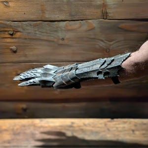 以下が含まれることがあります： 質感のある鱗模様の銀色の金属製のガントレットのペア。ガントレットは人の腕に着用されています。