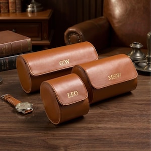 Könnte beinhalten: Drei braune Leder-Uhrenetuis mit personalisierten goldenen Initialen. Die Etuis sind zylindrisch mit einem Klappenverschluss. Eine braune Lederuhr mit silbernem Zifferblatt befindet sich neben den Etuis. Die Etuis tragen die Initialen "GW", "MHW" und "LEO".