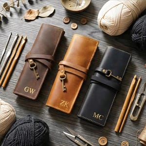 Puede incluir: Tres estuches de ganchillo de cuero en marrón, beige y negro, cada uno con una correa y un cierre en forma de llave. Los estuches están personalizados con iniciales. Ganchillos, hilo y otros suministros de artesanía están dispuestos alrededor de los estuches.