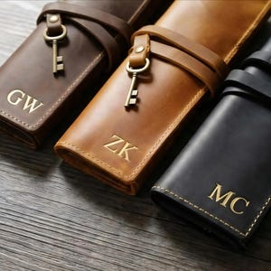 Puede incluir: Primer plano de tres estuches de cuero para bolígrafos en marrón, beige y negro. Cada estuche tiene una correa con un dije de llave y las iniciales doradas: "GW", "ZK" y "MC". Sobre una superficie de madera.