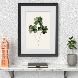 Pode incluir: Uma imagem emoldurada em preto com uma pintura em aquarela de tr&ecirc;s trevos de quatro folhas verdes. A imagem est&aacute; pendurada em uma parede branca texturizada acima de uma prateleira de m&aacute;rmore com um pequeno rel&oacute;gio Big Ben laranja, um livro preto e uma pequena planta em vaso.
