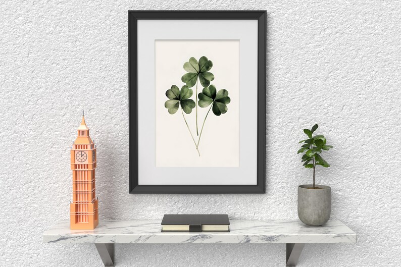Pode incluir: Uma imagem emoldurada em preto com uma pintura em aquarela de tr&ecirc;s trevos de quatro folhas verdes. A imagem est&aacute; pendurada em uma parede branca texturizada acima de uma prateleira de m&aacute;rmore com um pequeno rel&oacute;gio Big Ben laranja, um livro preto e uma pequena planta em vaso.