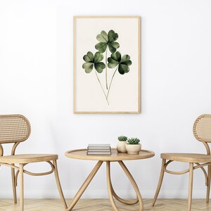 Pode incluir: Duas cadeiras de madeira clara com assentos e encostos de vime tecido, frente a frente em torno de uma mesa redonda de madeira clara com uma pilha de livros e duas pequenas plantas em vaso. Uma impress&atilde;o emoldurada de uma pintura em aquarela de uma planta de trevo de quatro folhas verde pendura na parede branca atr&aacute;s da mesa.