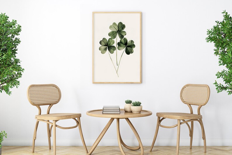 Pode incluir: Duas cadeiras de madeira clara com assentos e encostos de vime tecido, frente a frente em torno de uma mesa redonda de madeira clara com uma pilha de livros e duas pequenas plantas em vaso. Uma impress&atilde;o emoldurada de uma pintura em aquarela de uma planta de trevo de quatro folhas verde pendura na parede branca atr&aacute;s da mesa.