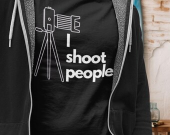 May include: Black t-shirt with a white graphic of a camera on a tripod and the text "I shoot people." The shirt is part of a photography-themed collection. The model is wearing a black zip-up hoodie.