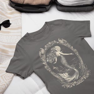 May include: A charcoal gray t-shirt with a vintage illustration of a mermaid in a decorative frame. The mermaid is depicted with long flowing hair and a tail, surrounded by floral details.