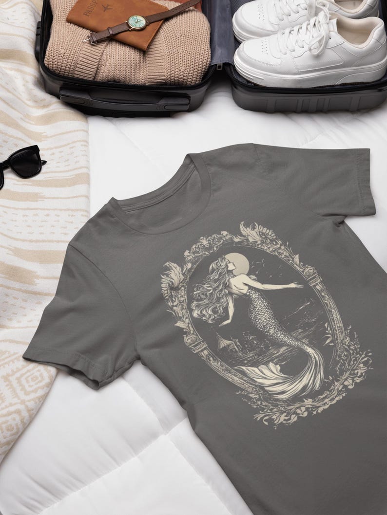 May include: A charcoal gray t-shirt with a vintage illustration of a mermaid in a decorative frame. The mermaid is depicted with long flowing hair and a tail, surrounded by floral details.