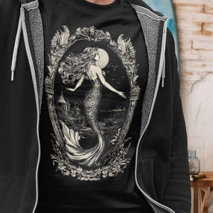 May include: A black t-shirt with a white graphic of a mermaid in an ornate frame. The mermaid is swimming in the ocean with a full moon in the background.