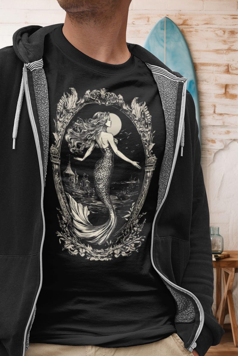 May include: A black t-shirt with a white graphic of a mermaid in an ornate frame. The mermaid is swimming in the ocean with a full moon in the background.