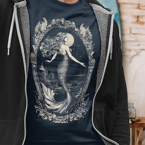May include: A navy blue t-shirt with a white graphic of a mermaid in an ornate frame. The mermaid is swimming in the ocean with a full moon in the background.
