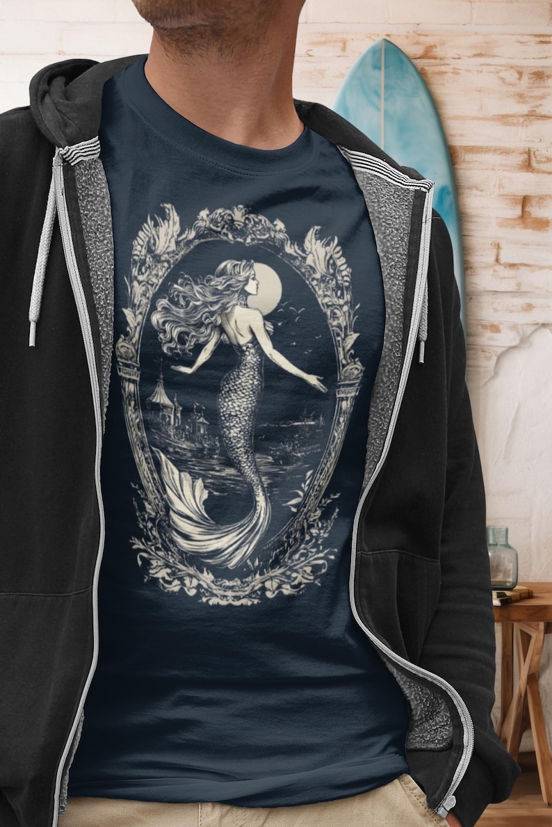 May include: A navy blue t-shirt with a white graphic of a mermaid in an ornate frame. The mermaid is swimming in the ocean with a full moon in the background.
