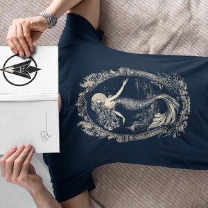 May include: A navy blue t-shirt with a white graphic of a mermaid in a decorative frame. The mermaid is depicted with long flowing hair and a tail.