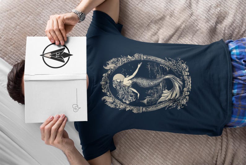 May include: A navy blue t-shirt with a white graphic of a mermaid in a decorative frame. The mermaid is depicted with long flowing hair and a tail.