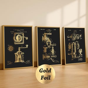 Könnte beinhalten: Drei gerahmte Patentdrucke mit goldfarbenen Illustrationen von Kaffeemühlen und Maschinen auf schwarzem Hintergrund. Die Drucke sind in goldenen Rahmen und werden auf einer Holzoberfläche ausgestellt.